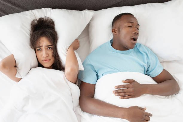 Snoring man sleeping with apnea and sleepless woman, angry wife unable to get to sleep. Upset young african american lady covers ears with pillow so as not to hear noise from snoring guy in bedroom
